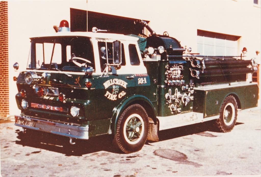 A vintage green fire truck with white accents from the Hillcrest Fire Co. is parked outside a building. The truck is equipped with extending ladders on the side and various hoses and valves.