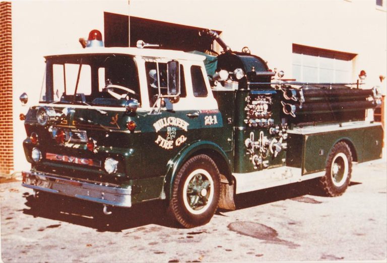 A vintage green fire truck with white accents from the Hillcrest Fire Co. is parked outside a building. The truck is equipped with extending ladders on the side and various hoses and valves.