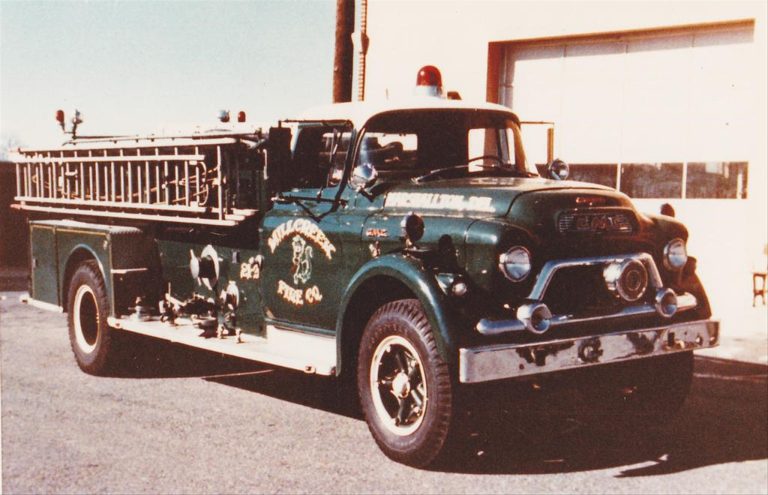 A vintage green fire truck with a chrome front bumper and grille is parked outside a fire station. It has a ladder on the side, a large hose reel, and emergency lights on top. The truck bears the name "Millcreek Fire Co." and appears to be well-maintained.