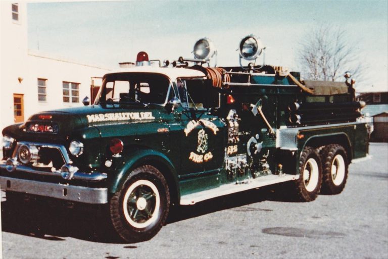 A vintage green fire truck with the label "Marshallville Fire Co." parked outdoors. The truck has large, round headlights, side-mounted tools, and hoses on top. The background shows a light-colored building and a leafless tree.