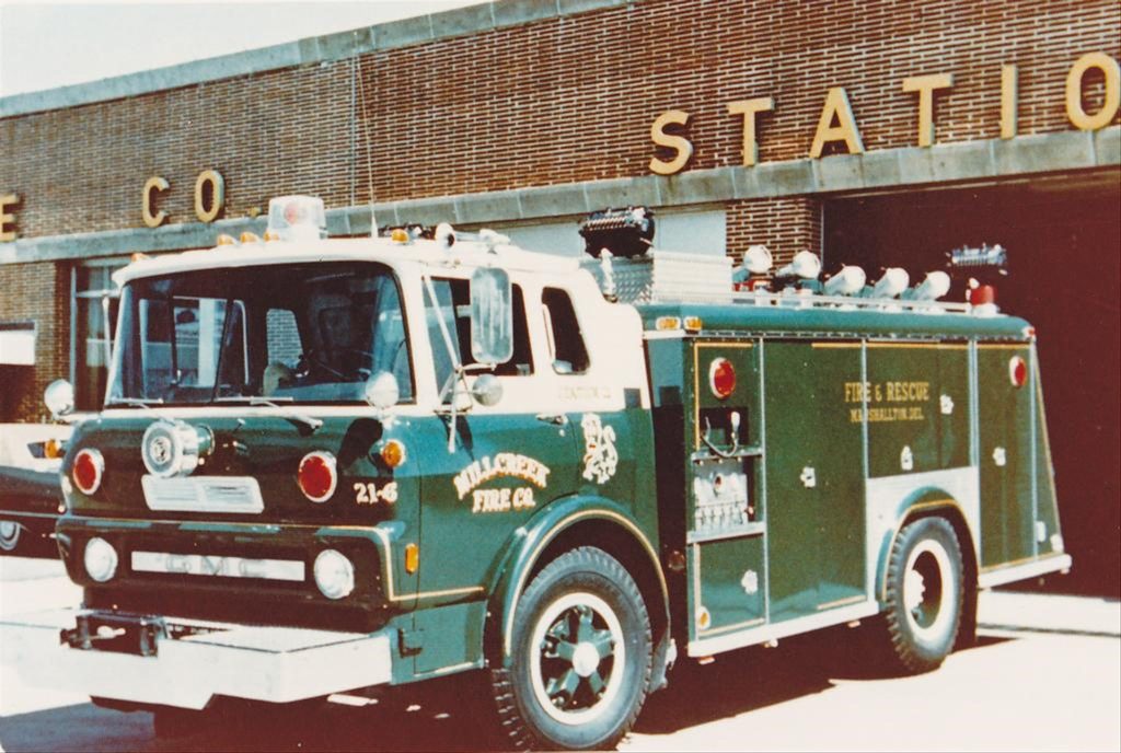 A green and white fire truck is parked in front of a fire station with the words "FIRE CO STATION" on the building. The truck has "Millcreek Fire Co." and emergency equipment on its side.