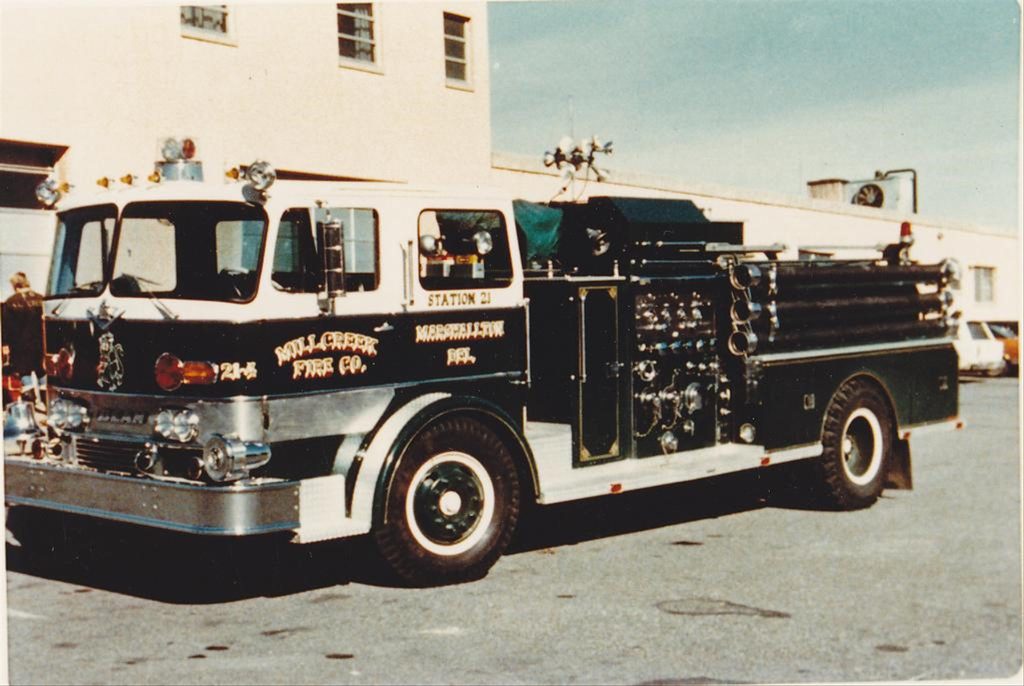 A vintage fire truck from Millcreek Fire Co. Station 21 is parked outside a building. The truck is dark green with silver detailing and various firefighting equipment compartments. It appears to be from the mid-20th century.