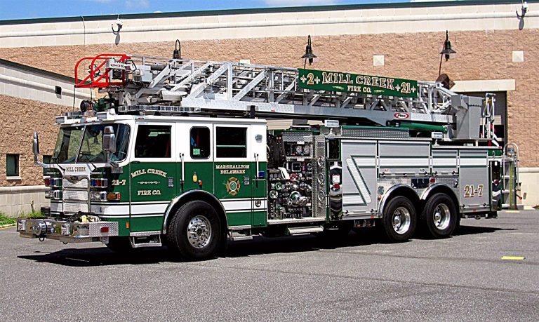 A green firefighting truck with "Mill Creek Fire Co." and "24-1" markings is parked outside a building. Its ladders and equipment are visible, and the truck has multiple storage compartments and water hoses. The sky is clear and the ground is paved.
