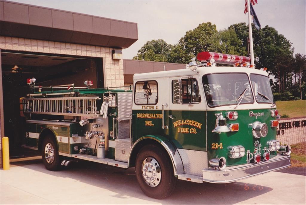 A green and white fire truck labeled "Marshalls Creek Fire Co." is parked outside a fire station. The truck has shiny chrome details, various equipment, and a roof-mounted light bar. The station has its door open, revealing the interior. Trees and a flagpole are visible in the background.