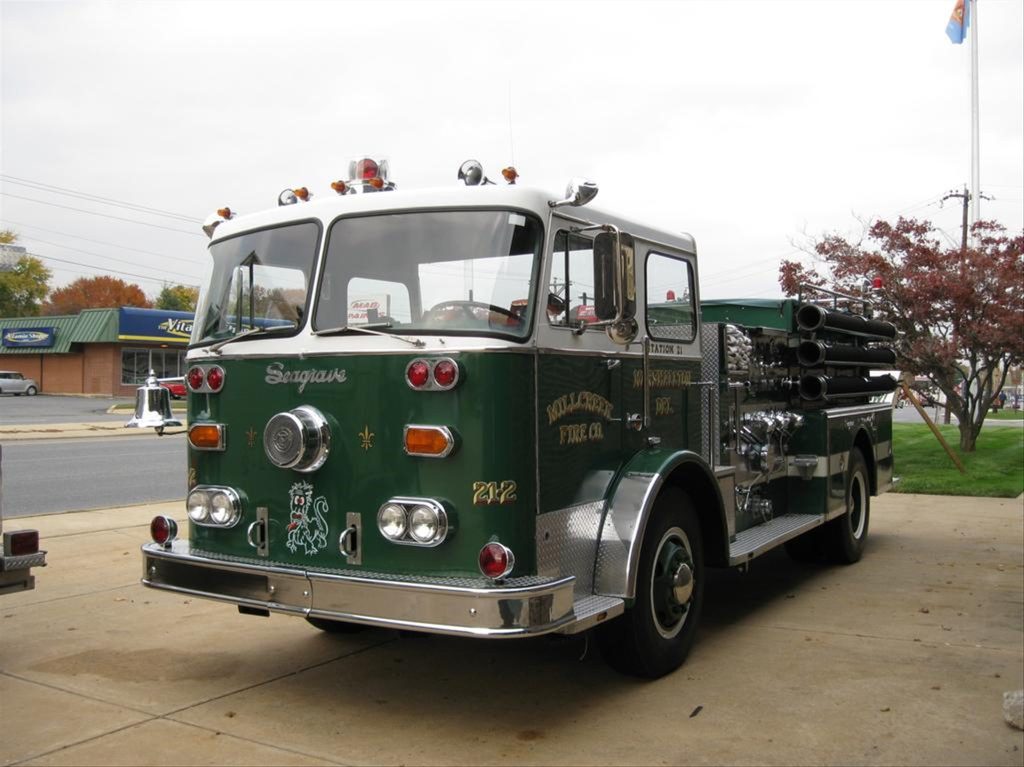 A vintage green fire truck is parked on a concrete driveway. The truck has '1960' written on it, along with text that reads "Salisbury Fire Co." and "Station 82." It has chrome accents, side mirrors, and multiple hoses on top. A small tree and buildings are in the background.