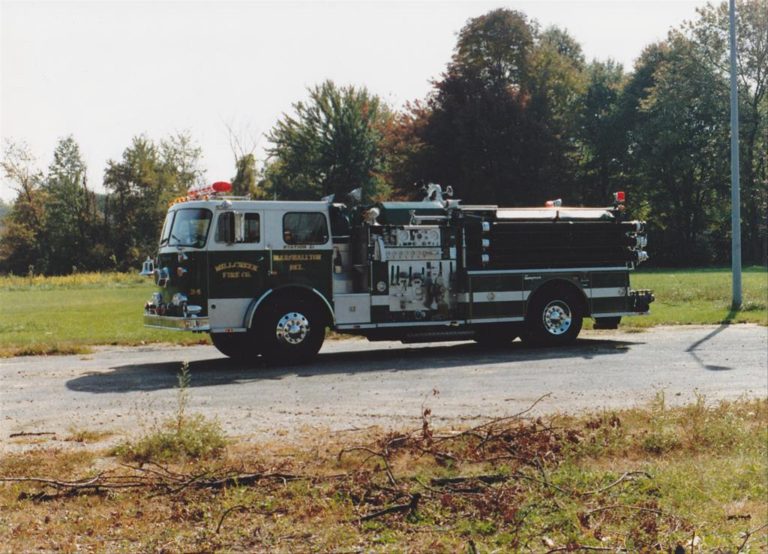 A fire truck with "Hillcrestville Fire Co." written on the side is parked on a paved area. The vehicle is equipped with various firefighting tools and has emergency lights on top. In the background, there is grass and a line of trees under a clear sky.