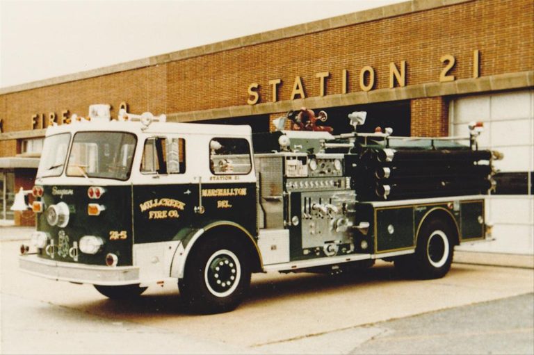 A vintage fire truck marked "Miller & Sons Fire Co.," labeled "Station 21" is parked in front of a brick building with "STATION 21" sign above large garage doors. The truck is green with chrome details and various firefighting equipment.