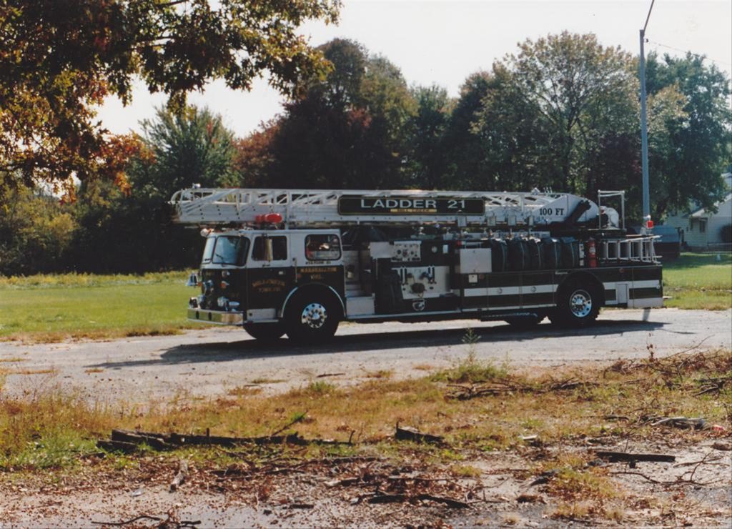 A fire truck labeled "Ladder 21" is parked on a paved area near a grassy field with trees in the background. The truck's ladder extends upward, reaching an impressive height. The scene captures a calm, sunny day with early autumn foliage visible on the trees.