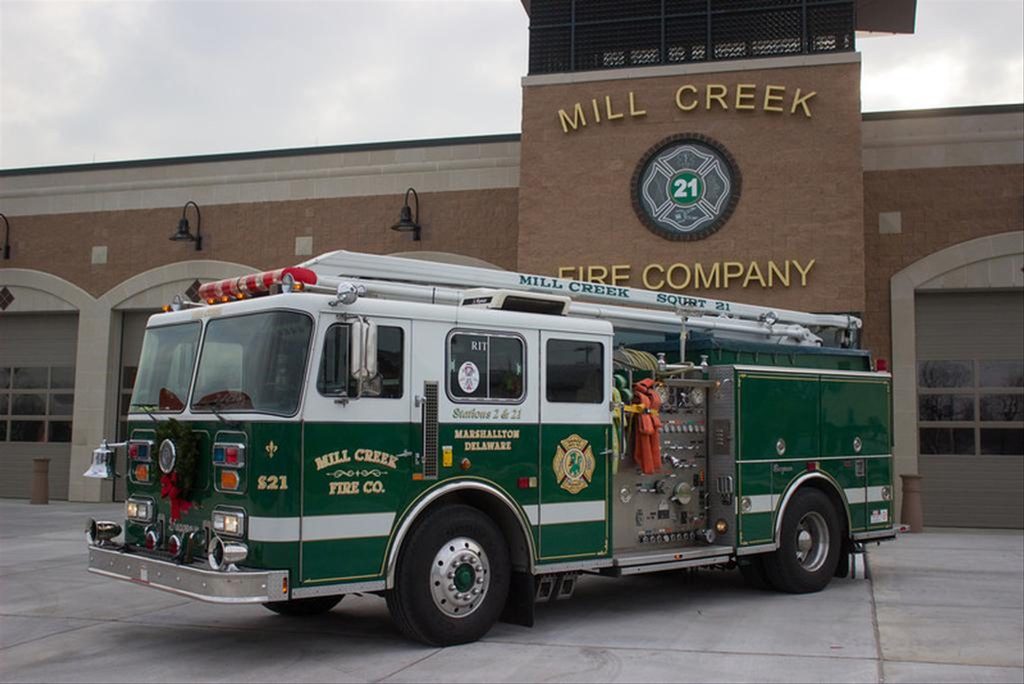 A green and white fire truck is parked outside the Mill Creek Fire Company station. The truck features various equipment and hoses. The station has three large garage doors, and "Mill Creek Fire Company" is displayed above the central entrance.