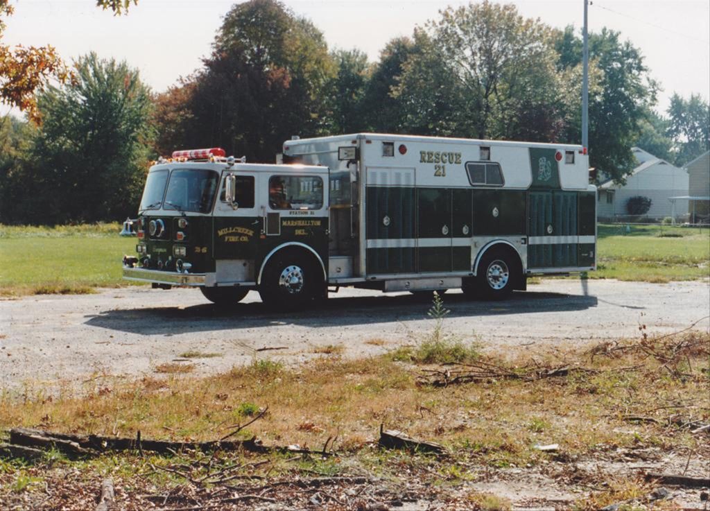A green and white fire rescue truck labeled "Rescue 21" is parked on a paved area, with houses and trees in the background. The truck has emergency lights on top and is part of the Naplate/Wallace FD, with "Marshallville" written on it.