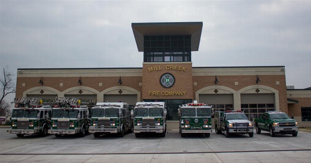 A lineup of fire trucks and an SUV parked in front of the Mill Creek Fire Company station. The building is brick with a large central tower and multiple garage doors. The sky is overcast.