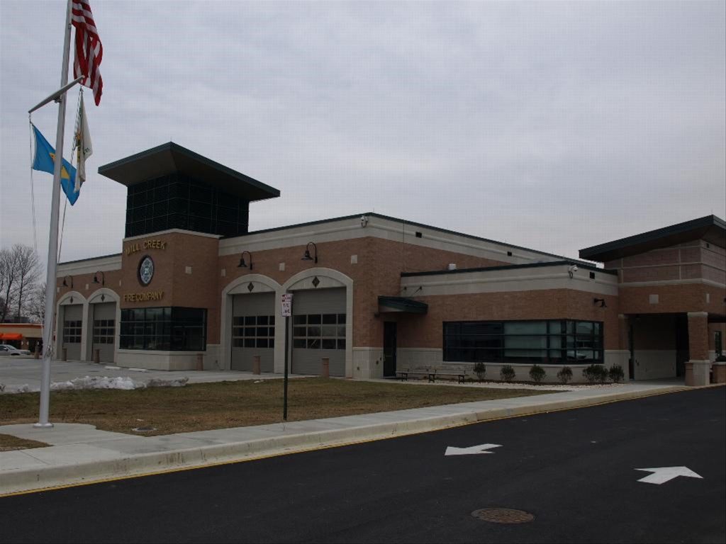 A modern fire station with a brick facade, featuring three bay doors. The building is flanked by a flagpole with three flags. The sky is overcast, and the area is surrounded by a paved road and sidewalk.