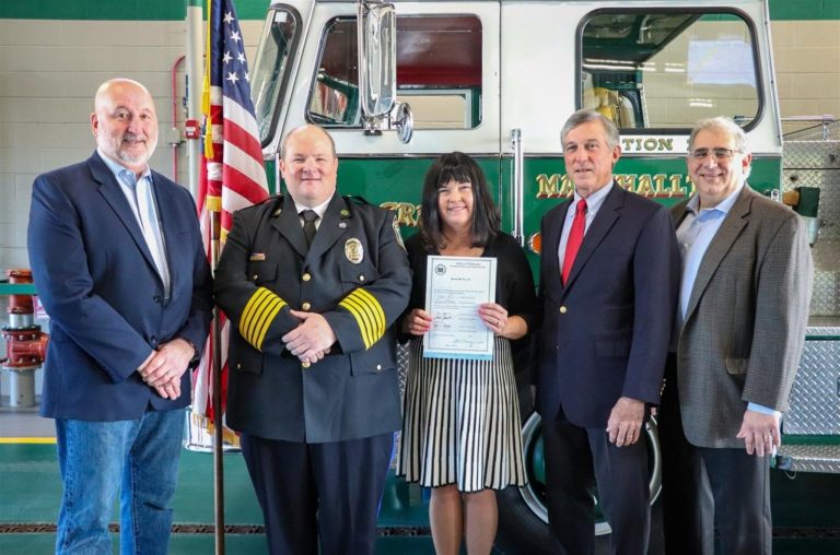 Five people stand in front of a green and white fire truck. A woman in the center holds a certificate. The group includes individuals in formal and fire department attire. An American flag is displayed nearby.
