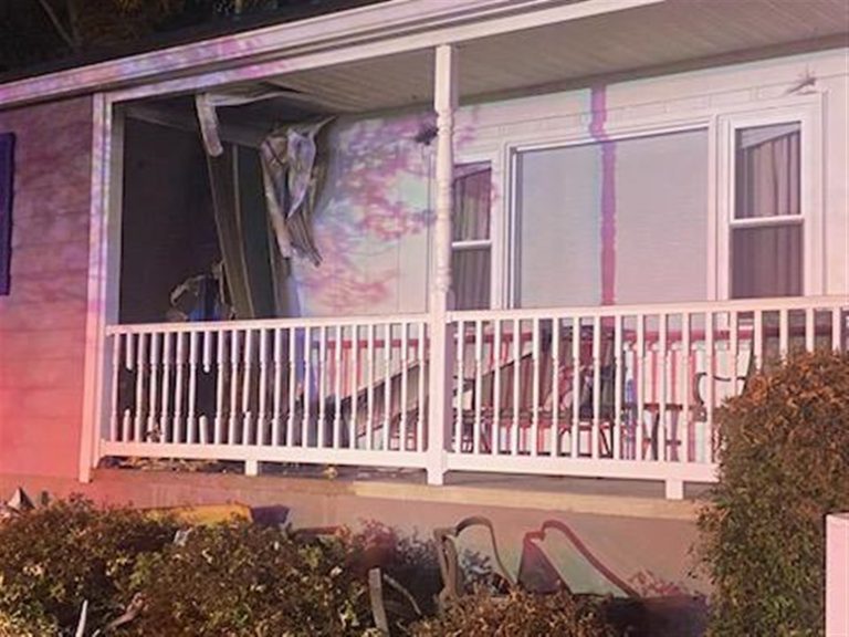 A house with structural damage is shown. The exterior wall and railing of the front porch are heavily damaged, including a large hole. Debris is scattered around the porch area. There are colorful lights illuminating the scene.