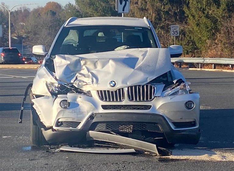 A silver SUV with significant front-end damage is stopped at an intersection. The hood is crumpled, and the bumper is detached. The background shows a road, trees, and a speed limit sign of 35 mph.