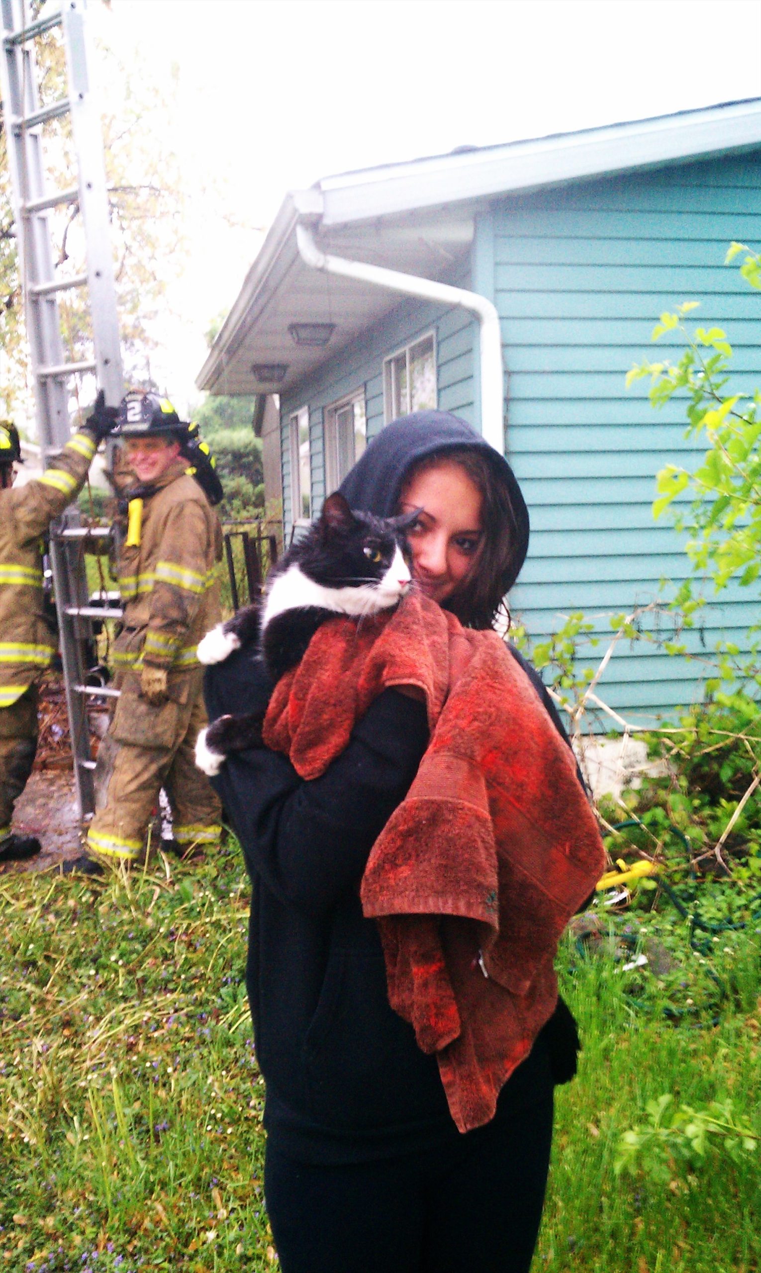 A person in a black hoodie holds a black and white cat wrapped in a brown towel outside a house. In the background, firefighters in uniforms are near a ladder leaning against the house. There is greenery around them.