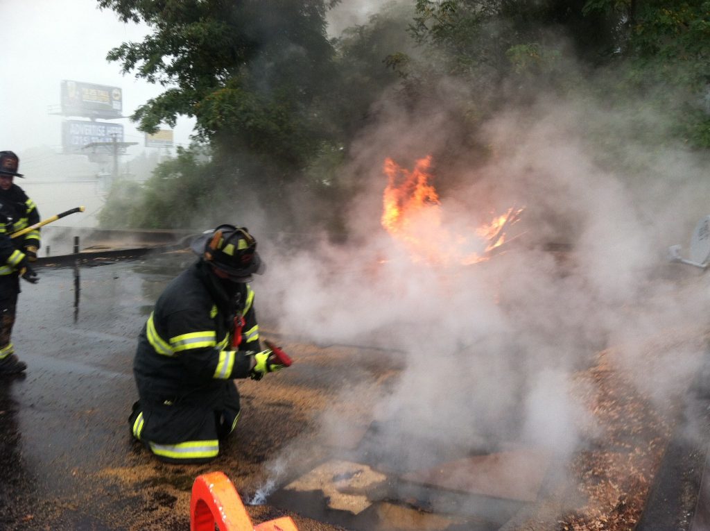 Firefighters in protective gear are on a rooftop surrounded by smoke, battling flames with hoses. A tree and a billboard are visible in the background. A firefighter kneels near the fire, working to extinguish it.