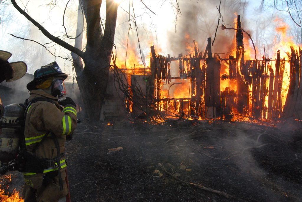 A firefighter in protective gear observes a blazing wooden structure engulfed in flames. The fire emits thick smoke, with trees silhouetted against the bright blaze in the background. Sunlight filters through the smoke and branches.