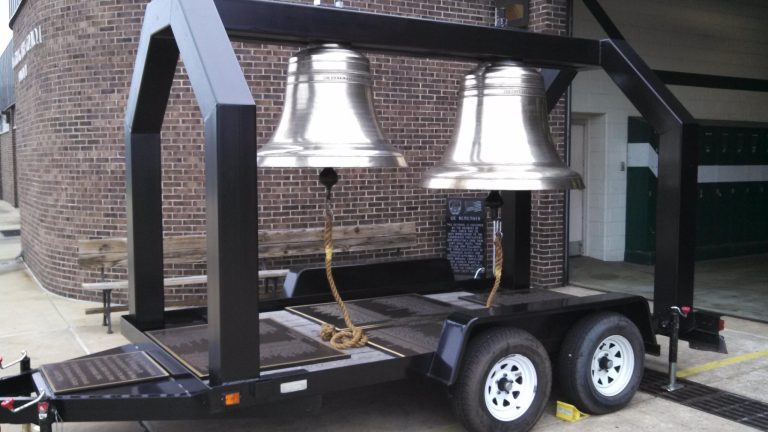 A mobile metal frame supports two large, polished silver bells on a trailer. The bells are secured with ropes. The trailer is parked on a concrete surface outside a brick building.