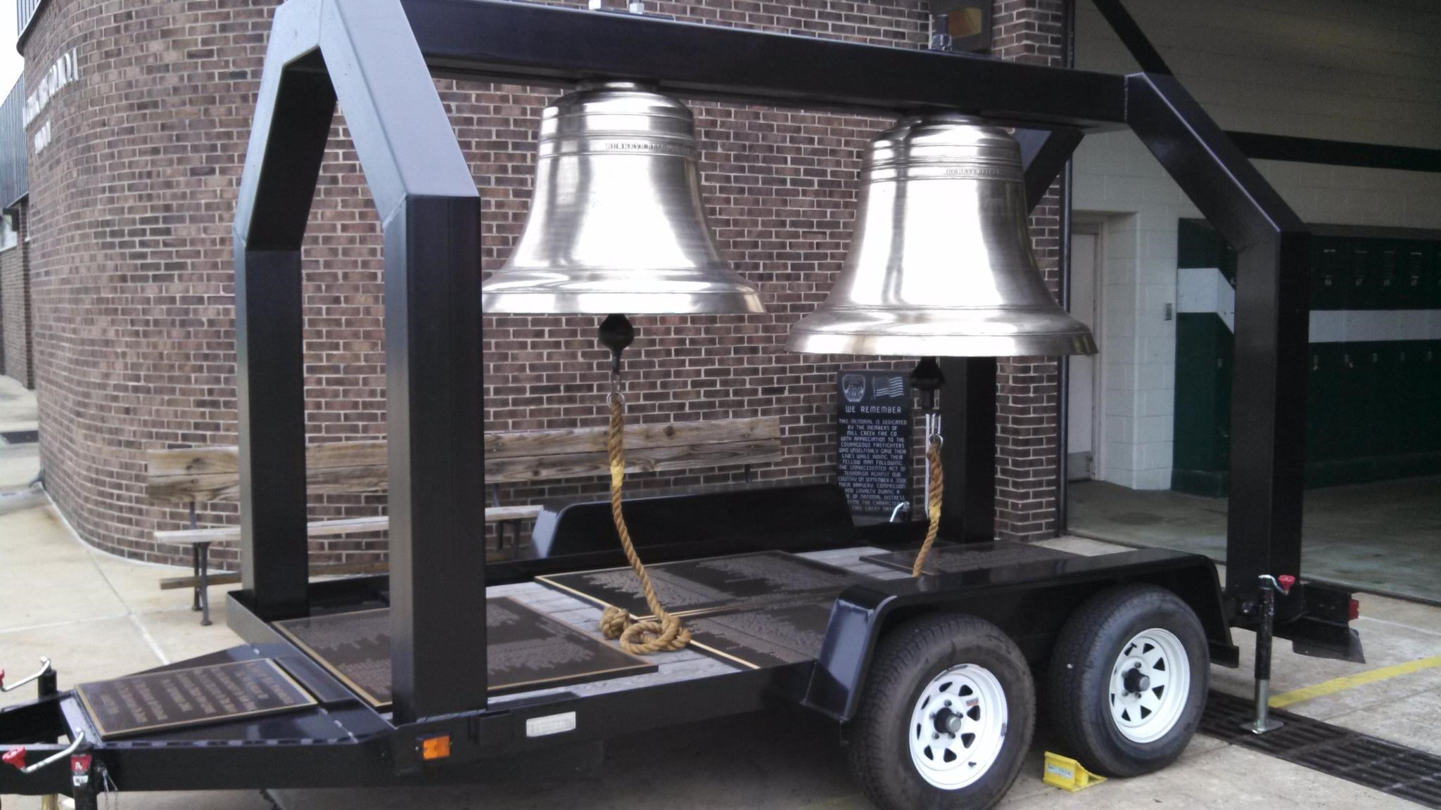 A mobile metal frame supports two large, polished silver bells on a trailer. The bells are secured with ropes. The trailer is parked on a concrete surface outside a brick building.