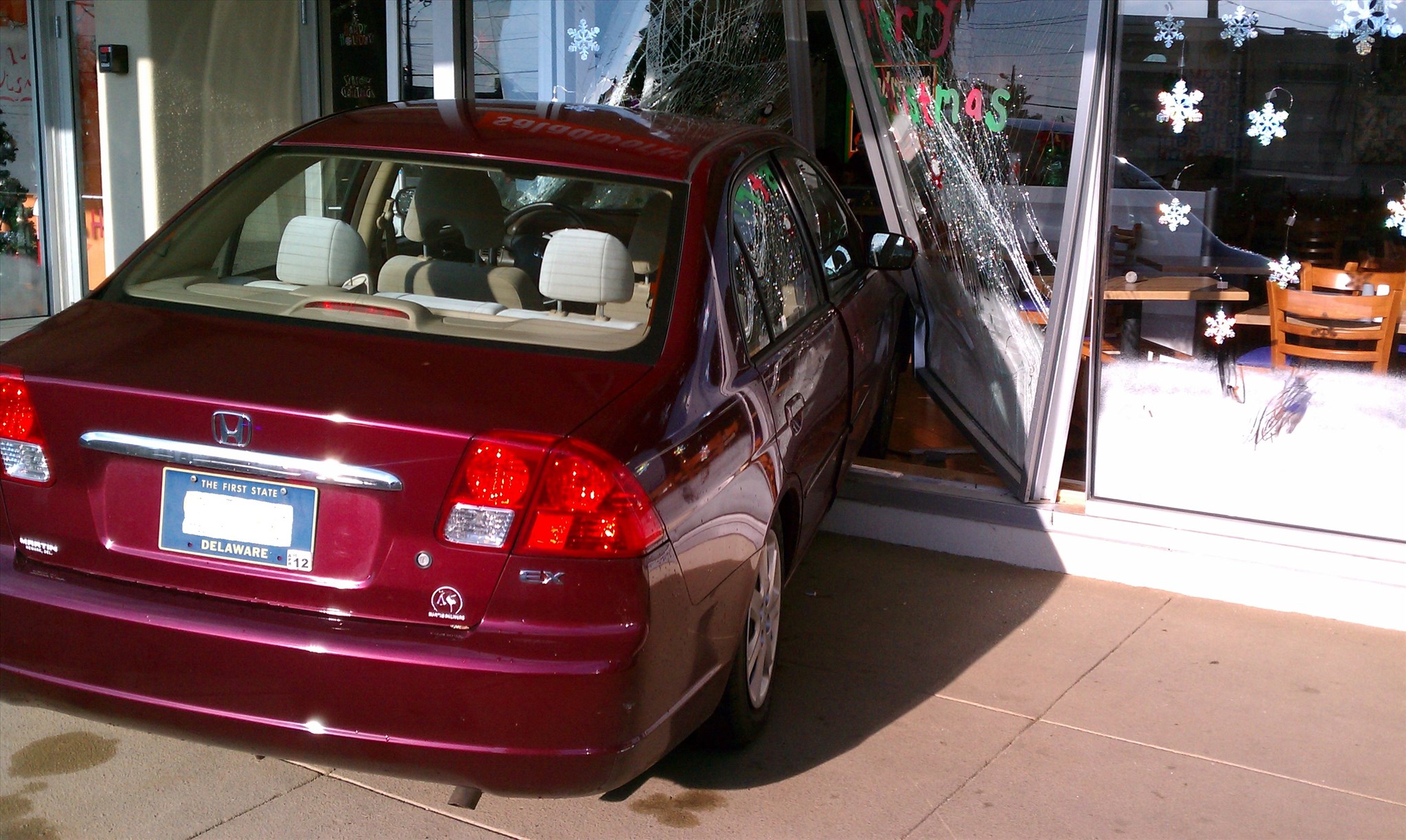 A maroon car has crashed into a building with large glass windows, causing significant cracks. Inside, chairs and tables are visible. Snowflake decorations are on the windows, suggesting a festive or holiday theme. The scene appears to be a restaurant.