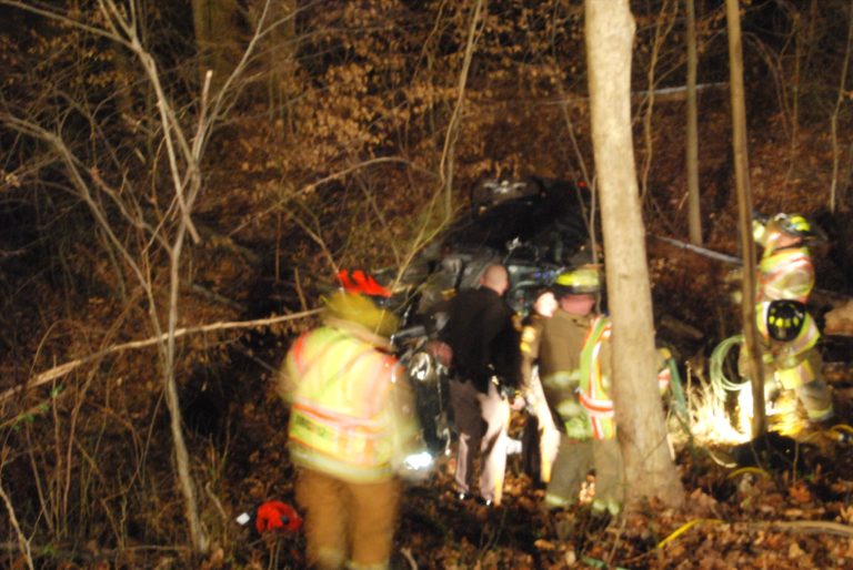 Firefighters and emergency responders are working at the scene of a car crash in a wooded area. The vehicle is partially visible among trees and debris. Responders in neon safety gear appear to be assisting at the site.
