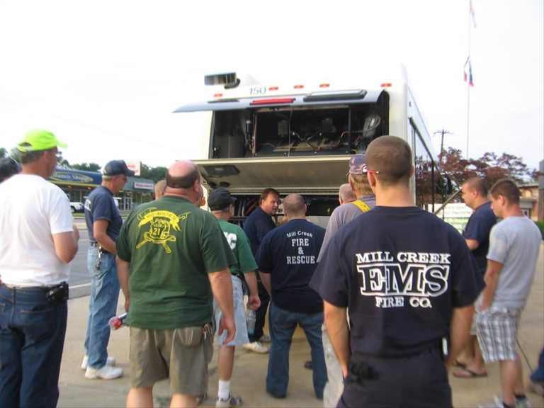 A group of people stand around the back of a large vehicle with its engine compartment open. They appear to be examining or discussing something inside. Some individuals are wearing shirts with "Fire & Rescue" and "EMS" on them.