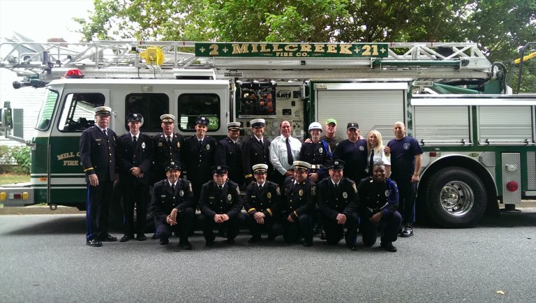 A group of firefighters and staff pose in uniform in front of a fire truck labeled "Mill Creek Fire Co. 21." The truck is green and silver. Trees and a road are visible in the background.