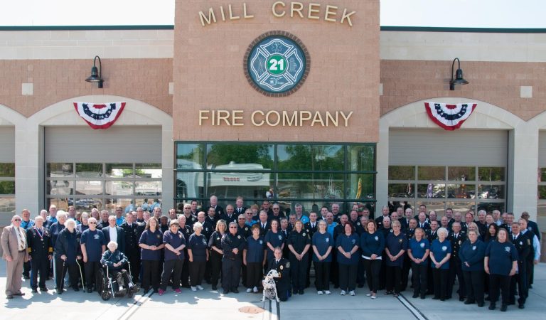 A large group of people stand in front of the Mill Creek Fire Company building. They are wearing matching navy blue uniforms. The building has garage doors and a sign with the number 21. Festive banners adorn the structure.