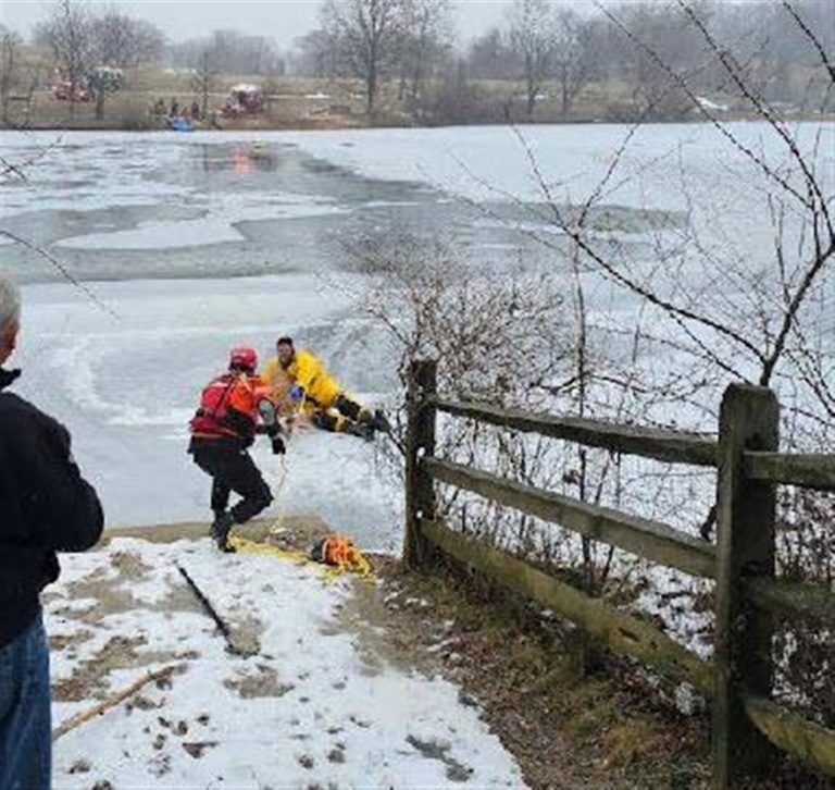 A rescue worker in a red outfit assists a person on the edge of a partially frozen lake. Another person stands nearby observing the scene. Snow covers the ground, and leafless trees are visible in the background.