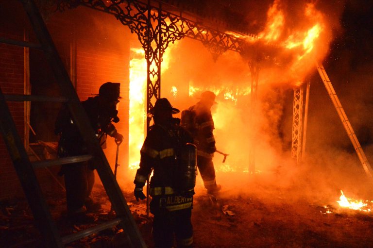 Firefighters in full gear work to extinguish intense flames engulfing a building. The scene is filled with smoke and fire, casting a dramatic orange glow. Ladders are visible on the sides, and ornate metalwork frames the porch area.