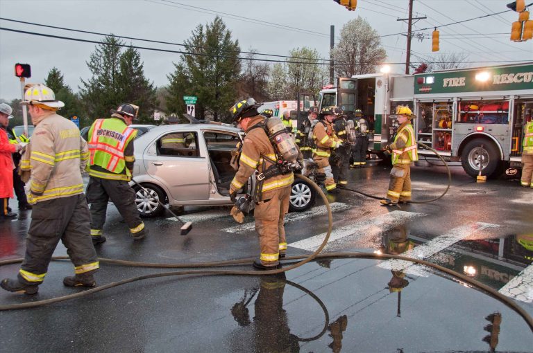 Firefighters in gear respond to a car accident at an intersection on a rainy day. They surround a silver vehicle with open doors, hoses on the wet road. A fire truck is parked nearby with flashing lights. Trees are visible in the background.