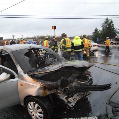 Firefighters in yellow and orange gear respond to a car accident on a wet road. A silver car with significant front-end damage is at the center, while emergency personnel work around the scene. Traffic lights and trees are visible in the background.