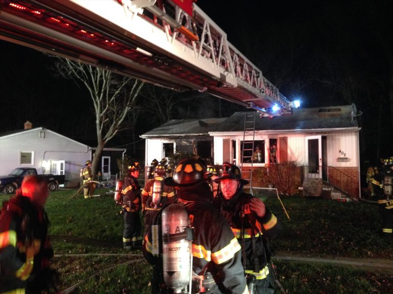 Firefighters in gear gather outside a small white house at night. A fire truck ladder extends toward the roof, and a ladder leans against the building. The scene is illuminated by emergency lights.