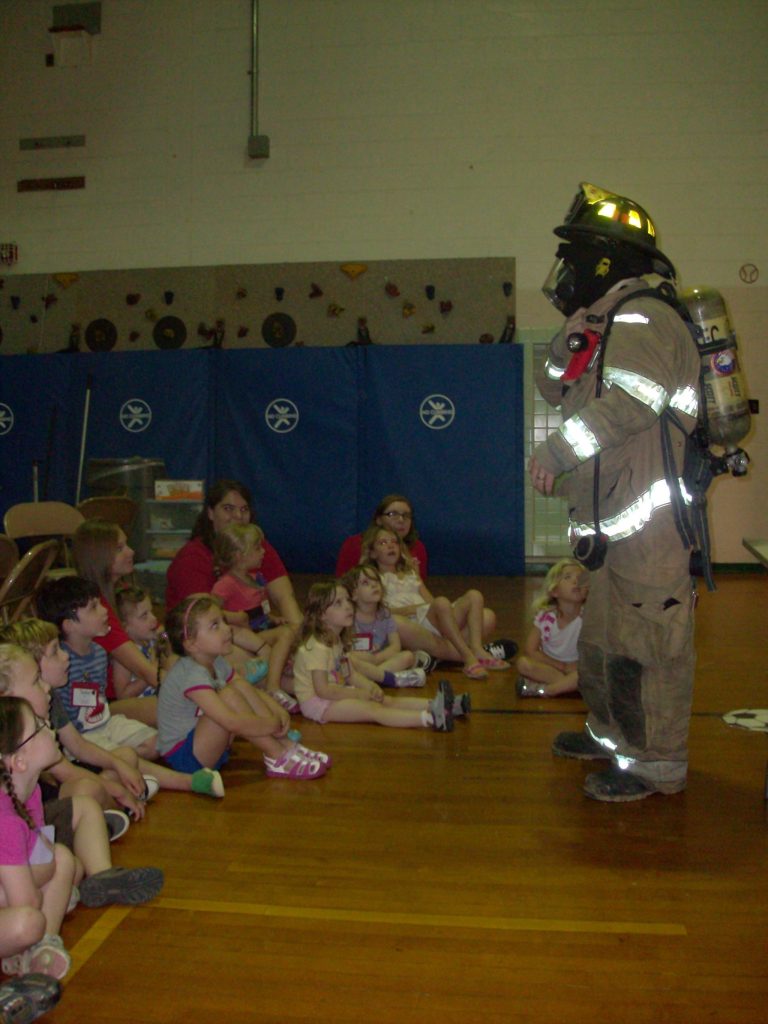 A firefighter in full gear stands in a gymnasium, speaking to a group of young children who are seated on the floor. The children listen attentively. The room has blue padded walls and climbing equipment in the background.