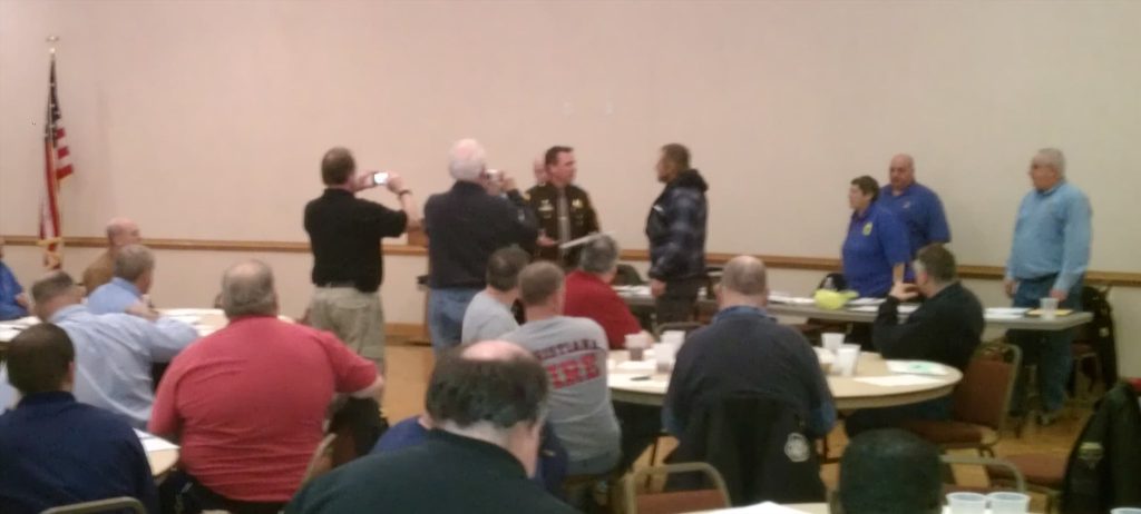 A group of people is in a room with tables and chairs. One person is being photographed by others in the center. An American flag is in the background. Attendees are seated, facing the front.