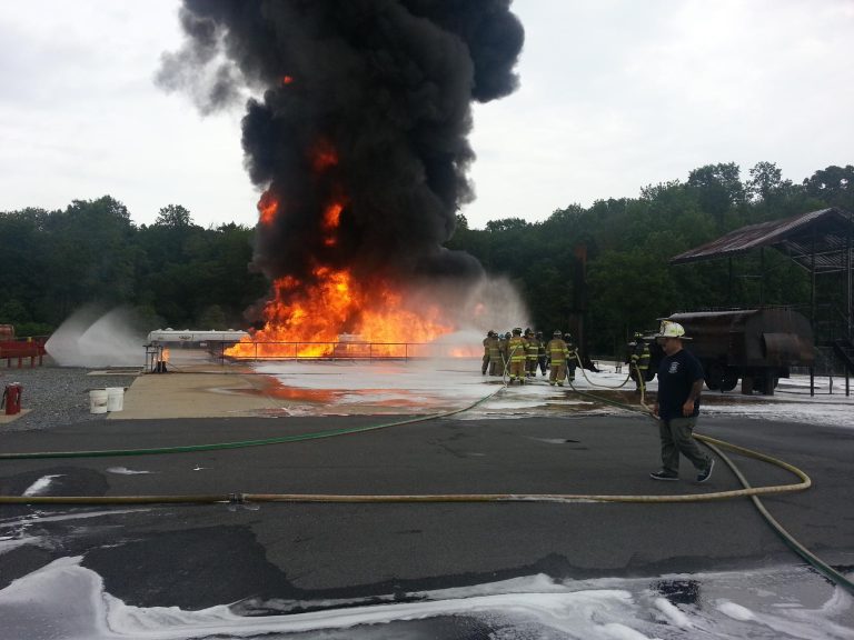 A group of firefighters is battling a large fire with thick black smoke billowing into the sky. They are using hoses to spray water on the flames, surrounded by trees. A firefighter walks in the foreground.