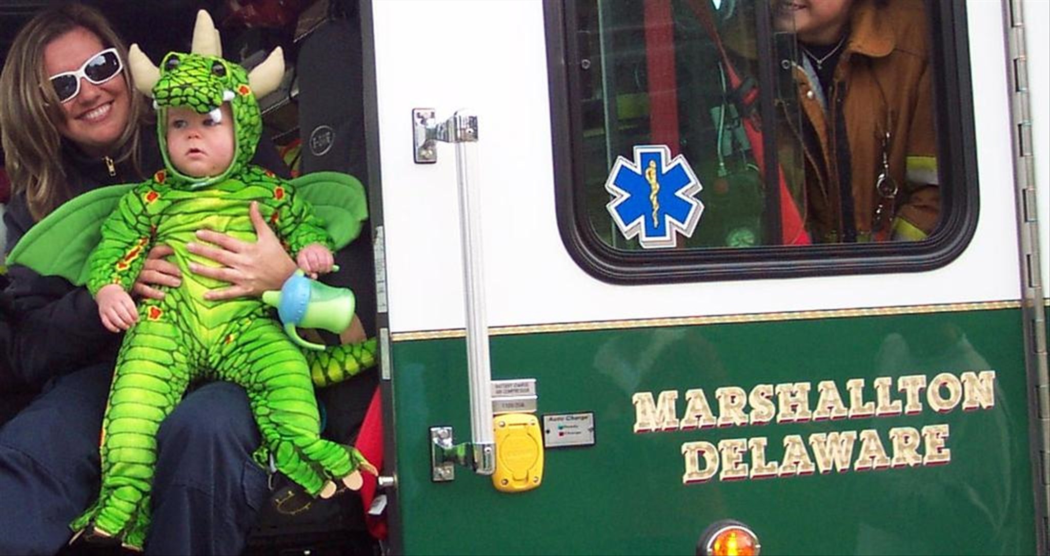 A woman holds a baby dressed in a green dragon costume, sitting in the open doorway of a Marshallton, Delaware fire truck. A firefighter is partially visible inside the truck. The fire truck door features an emergency medical symbol.