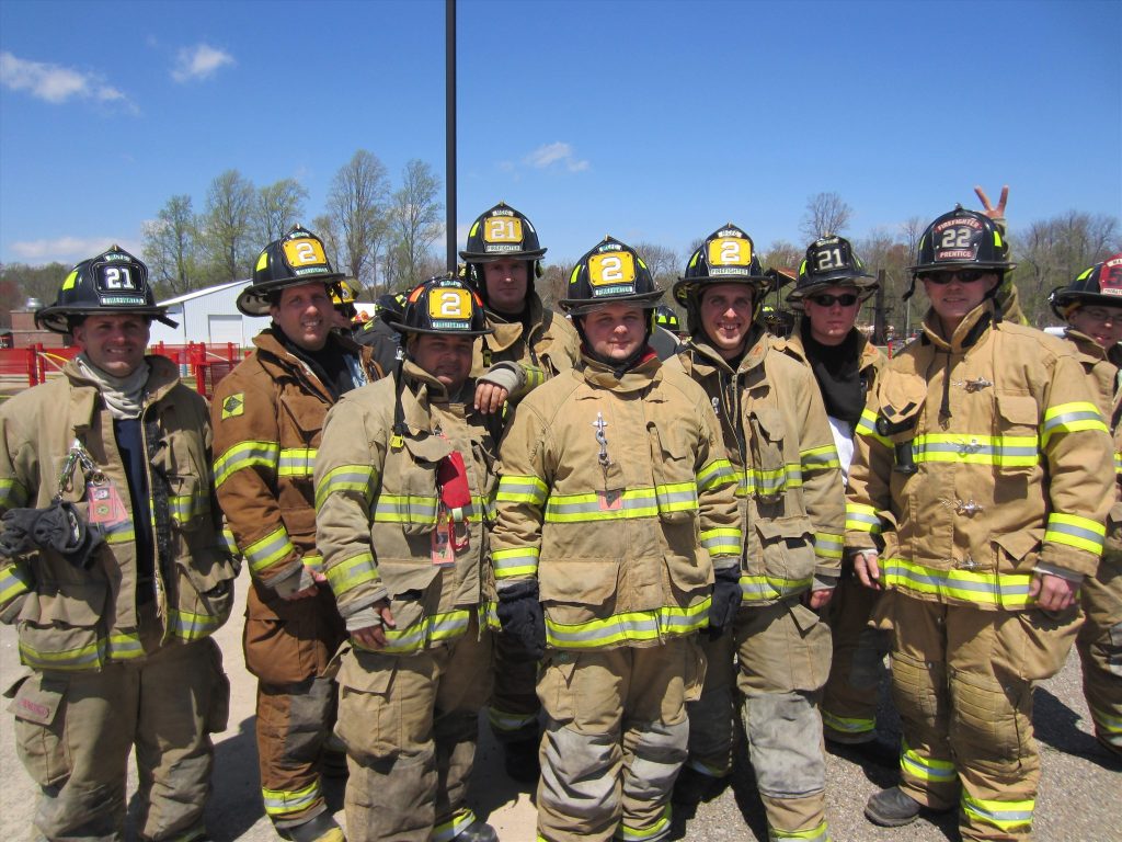 A group of firefighters wearing tan protective gear and helmets with numbers "21" and "22" pose outdoors. The sky is clear, and trees are visible in the background.