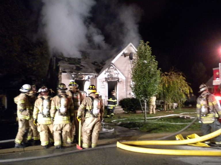 Firefighters in gear gather outside a house at night. Smoke billows from the roof, and a hose is laid out on the street. Emergency lights illuminate the scene, and trees are visible near the house.