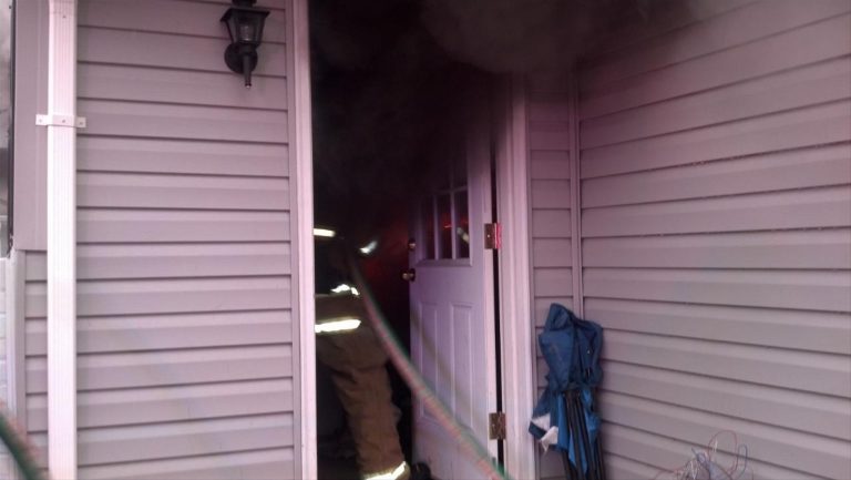 A firefighter wearing reflective gear enters a smoke-filled house through an open door. The building has gray siding, and an umbrella is propped near the entrance. Thick black smoke obscures the interior.