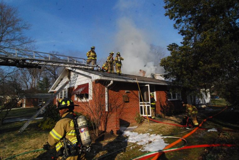 Firefighters in gear stand on the roof of a brick house, tackling smoke and a fire. A ladder extends to the roof, and hoses are spread across the lawn. Sparse trees and another house are visible in the background.