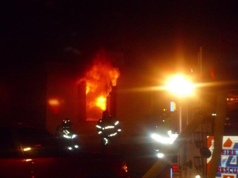 Firefighters respond to a building fire at night. Flames are visible through a window, and bright lights illuminate the scene as emergency personnel work to control the blaze.