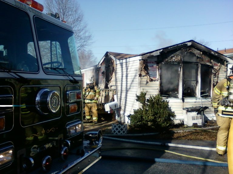 A firetruck is parked beside a damaged house with charred walls and a collapsed roof. Several firefighters in gear are assessing the situation, with hoses on the ground and smoke visible around the structure.
