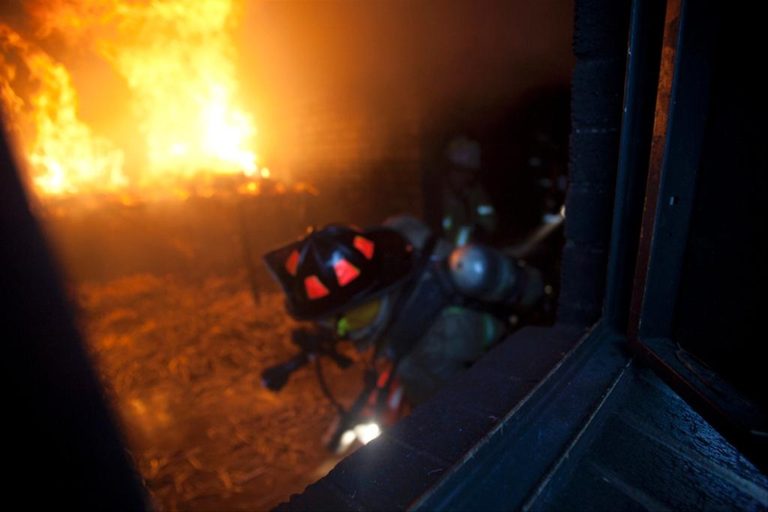A firefighter in gear crouches near a window in a burning building, with bright flames filling the background. The room is filled with smoke and debris, highlighting the intensity of the fire.