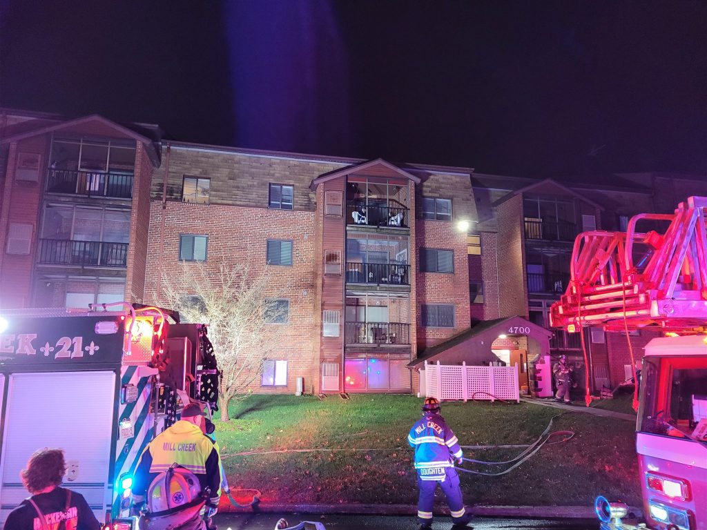 Firefighters respond to a nighttime emergency at a multi-story residential building. Firetrucks and emergency personnel are positioned on the street, with hoses and equipment in use. The building shows evidence of a fire in an upper-floor apartment.