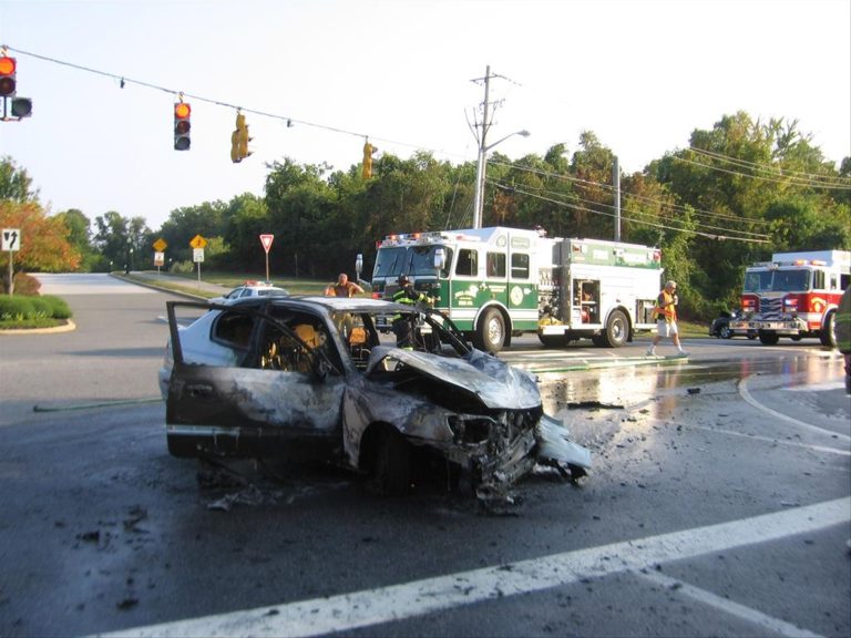 A charred car is seen at an intersection with firefighters and fire trucks nearby attending the scene. Debris is scattered around the vehicle. Traffic lights are visible in the background. Trees and clear skies form the backdrop.