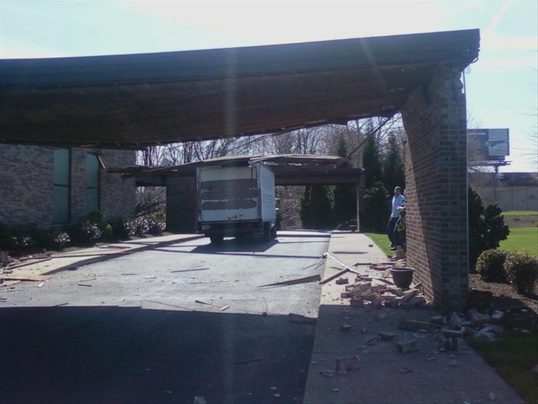A white box truck is lodged under a partially collapsed brick carport, with debris scattered on the ground. A person stands nearby, looking at the scene. The sky is clear and trees are visible in the background.