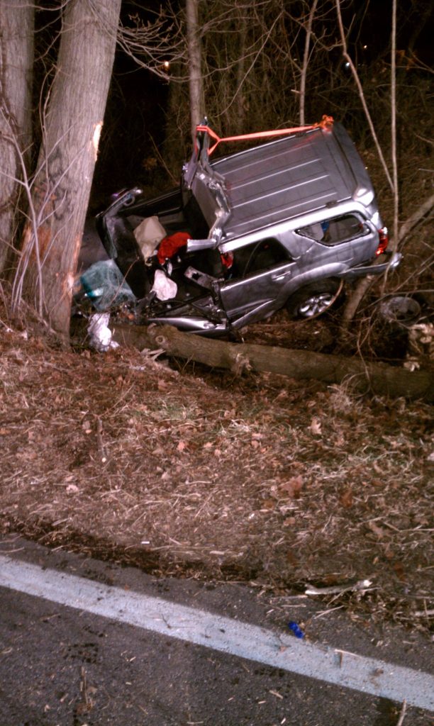 A silver SUV is crashed between two trees beside a road at night. The vehicle is severely damaged, with its rear lifted and trunk access open. The surrounding area is wooded, with scattered debris and dry leaves.