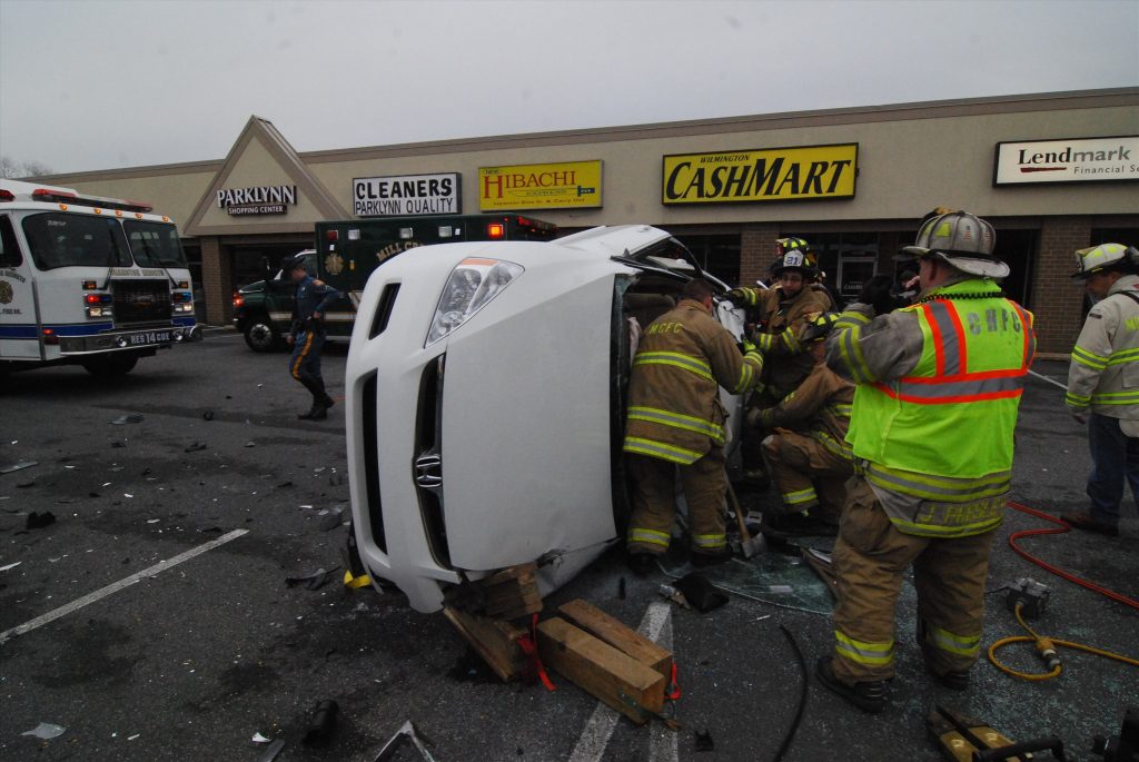 Firefighters work at the scene of an accident involving a white car flipped on its side in a parking lot. Nearby are emergency vehicles and several store fronts, including a cleaners and a restaurant. Debris is scattered on the ground.